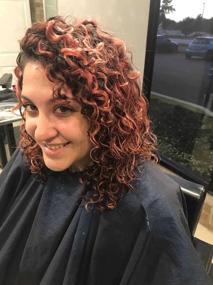Short Deva curly cut with copper-red highlights on shoulder-length curls, smiling client by Richie at Damron & Company Salon in Austin